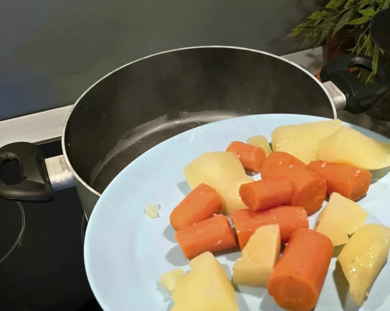 A blue plate with large chunks of boiled potatoes and carrots, being prepared to be added to a steaming pot on the stove.
