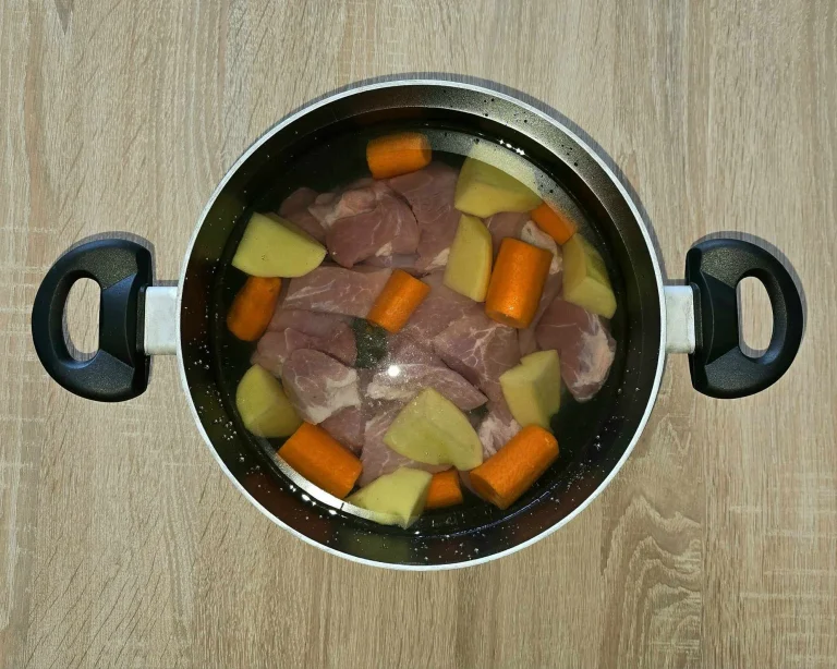 A top-down view of a pot filled with raw pork pieces, chopped carrots, and potatoes submerged in water, placed on a wooden surface, ready for cooking.