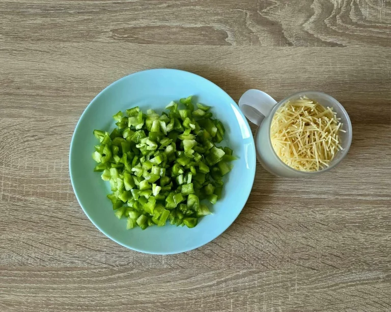 A blue plate with freshly chopped green bell peppers and a transparent mug filled with thin, broken noodles, placed on a wooden surface.
