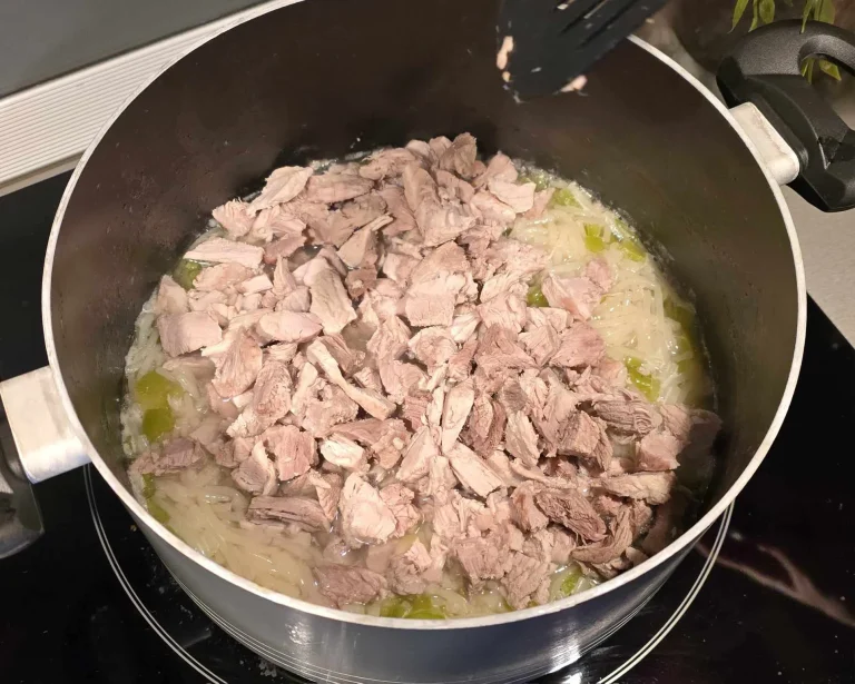 A pot of soup on the stove with shredded pork, noodles, and chopped green vegetables.
